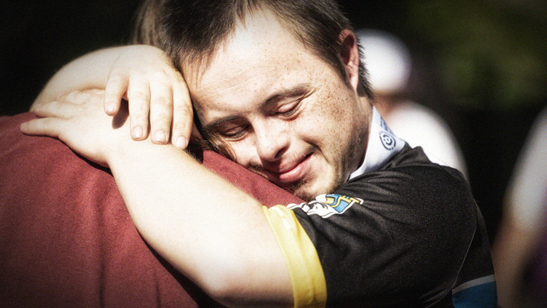 Ein Mann mit Down-Syndrom umarmt eine Person und lächelt mit geschlossenen Augen.