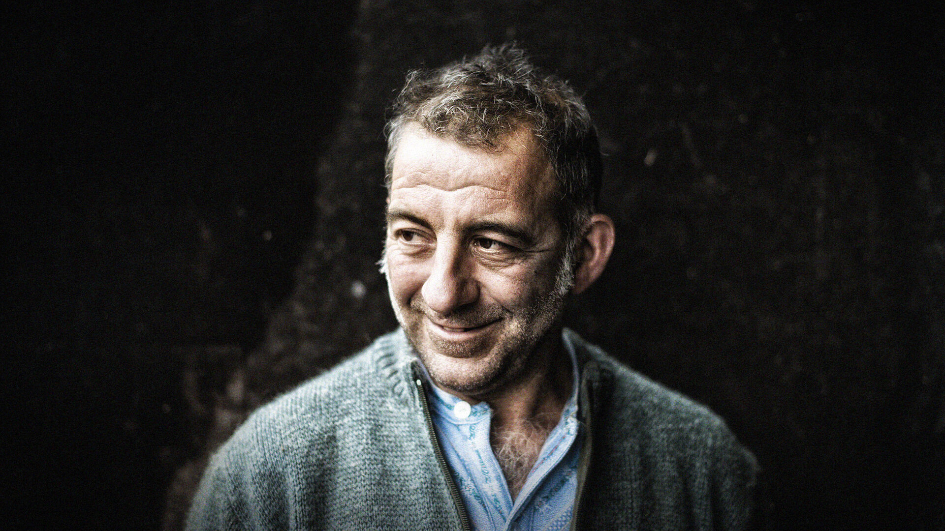 Lächelnder Mann in grauer Strickjacke und Hemd vor dunklem Hintergrund im Porträt.